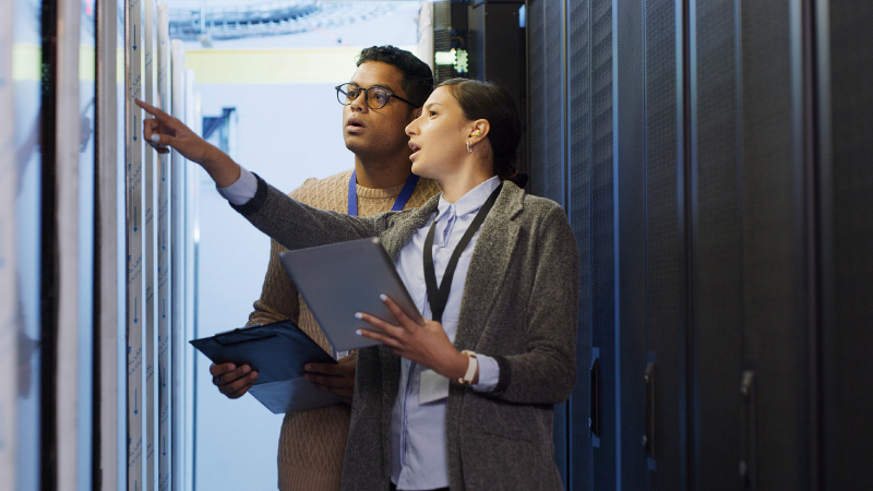 Two people in business attire are in a server room. One is pointing at a server rack while the other holds a tablet, both focused and engaged.