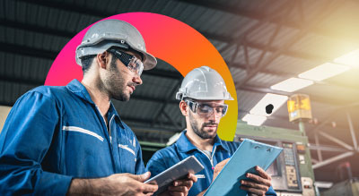 Two men wearing hard hats and protective glasses looking at tablet and clipboard in a factory