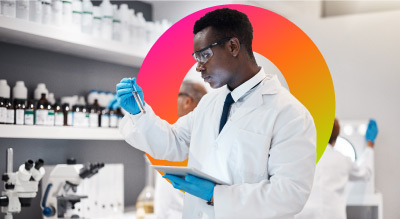 A scientist in a lab coat and gloves is examining a blue liquid in a test tube while holding a tablet, with colleagues working in the background