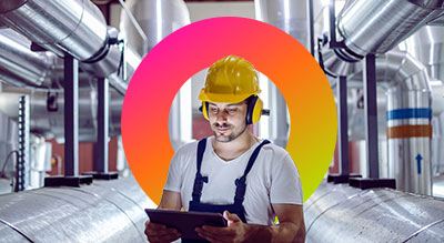 A worker in a hard hat and headphones holding a clipboard in a factory with pipes in the background