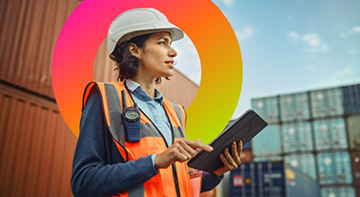 An inspector wearing a hardhat and high-visibility vest works on a digital tablet with freights in the background