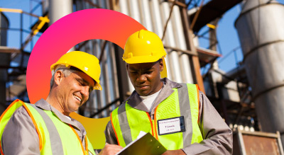 Two workers in safety vests and helmets discussing notes on a clipboard at an industrial site