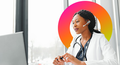 A healthcare professional with a stethoscope is seated in front of a laptop while wearing a headset