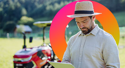 A person standing outdoors, wearing a light-colored shirt and a straw hat, holding a tablet or clipboard. Behind them, a red tractor is parked on a grassy field