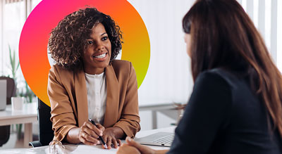 Two people in a work setting, one assisting the other with paperwork on a desk