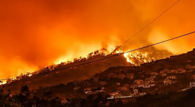 A devastating wildfire engulfing a hillside with houses, under an ominous orange sky
