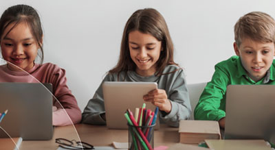 Three children use laptops and tablets in a classroom