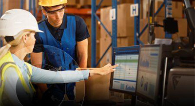 Two individuals wearing hard hats are working in a warehouse, one pointing at a computer screen while the other observes