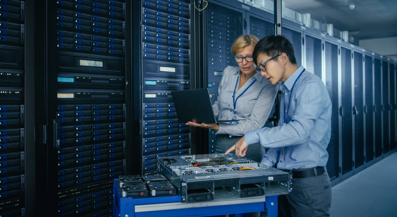 Two individuals in a modern server 
                  room with one holding a laptop