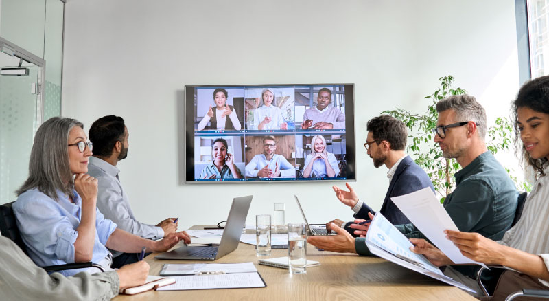 A group of people seated around a conference table participating in a video call, with a large monitor mounted on the wall displaying remote participants.