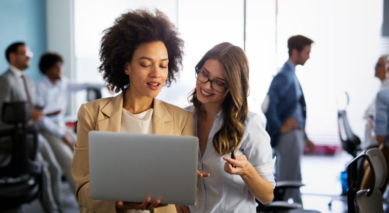 Two individuals standing in an office setting with people in the background while they look at a laptop
