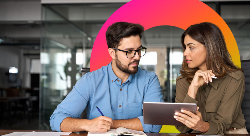 Two individuals are looking over a tablet while one is writing in a notebook