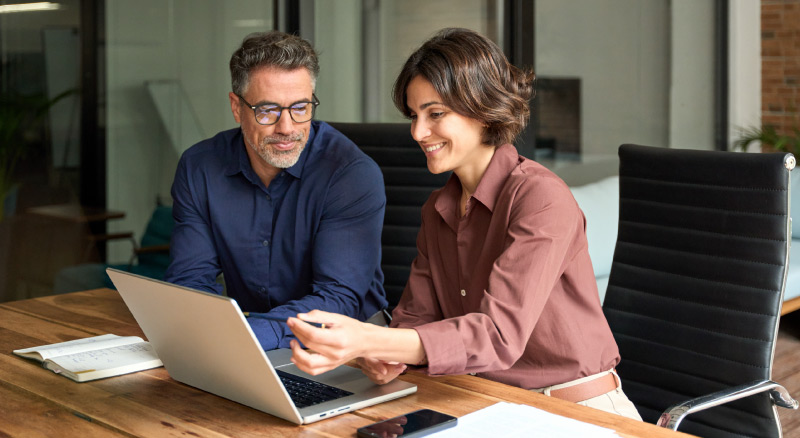 A smiling individual writes notes while using a laptop