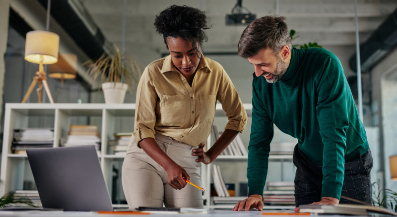A smiling individual writes notes while using a laptop