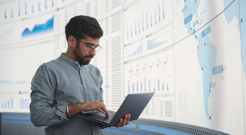 Employee analyzing information on a laptop while data is displayed on large screens in the background