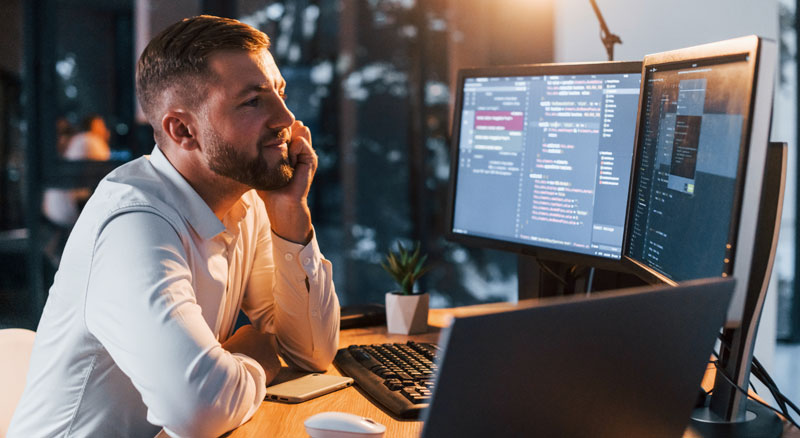 A person in a white shirt is coding at a desk with multiple computer monitors, illuminated by their glow