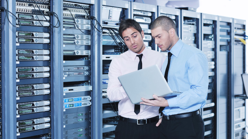 Two individuals stand in front of server racks while one holds an open laptop