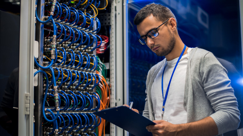 A person in front of a server rack, holding a clipboard, with numerous network cables organized in the background