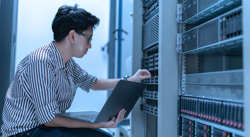 A person on a laptop in a server room inspecting the server equipment