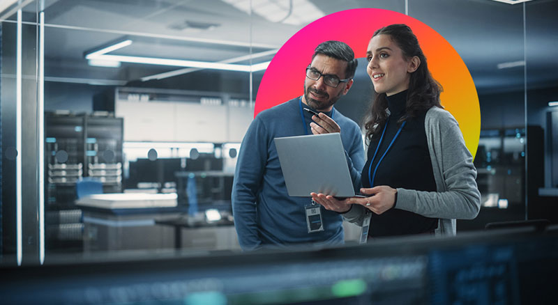 Two individuals stand in a modern server room with one holding a laptop