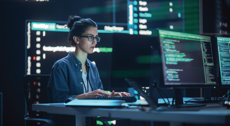 A person sitting in front of multiple computer monitors displaying lines of code, working in a dimly lit room