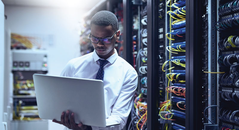 A person in a white shirt and tie holding a laptop while standing in front of server racks in a data center