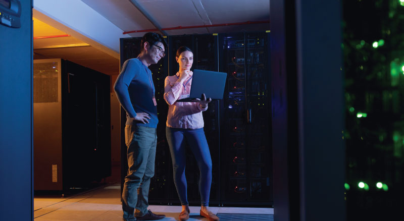 Two individuals in a server room, one standing with hands on hips looking at server racks, the other holding a laptop