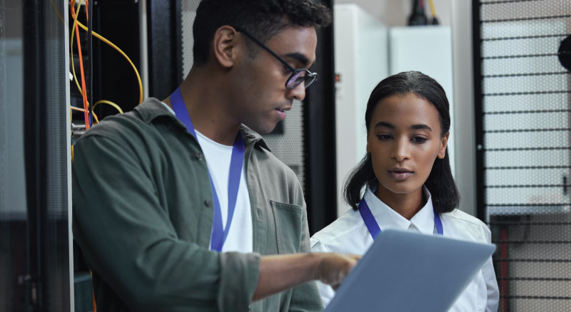 Two individuals standing in a server room, one holding a laptop and the other holding a clipboard, both wearing lanyards, seemingly engaged in work-related tasks