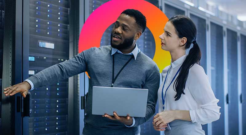 Two individuals stand before a server, one holding a laptop, engaged in a discussion about technology.