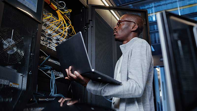 A person in a server room, holding a laptop and examining network equipment