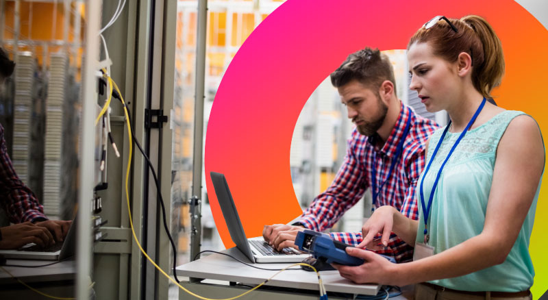 Two employees in a server room. One employee is on a laptop.