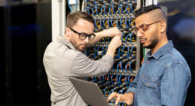 Two men in a server room. One is working on his laptop.