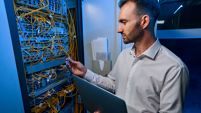 man adjusts wires in server room referring to a tablet