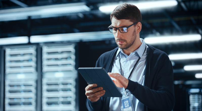 A person stands in a server room, holding and looking at a tablet