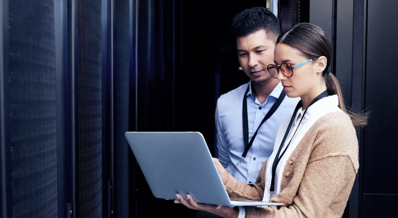 Two individuals review information on a laptop in a server room