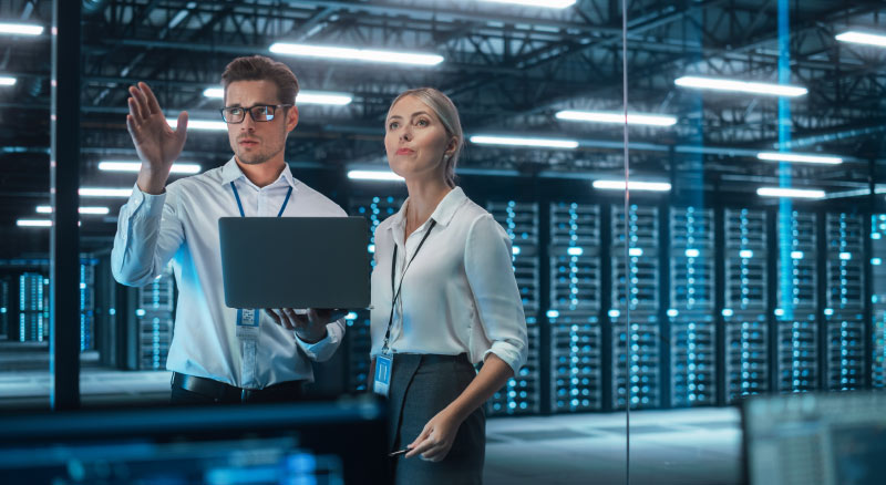 Two individuals stand in a server room with one holding holding a lapto