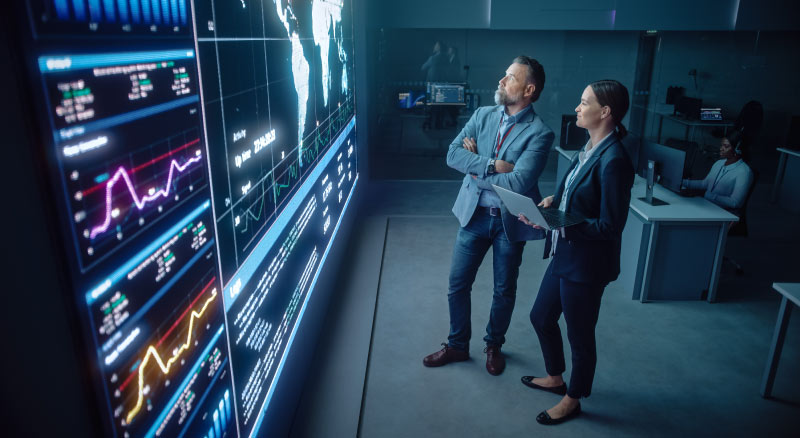 Two professionals are analyzing data on large digital screens in a high-tech control room