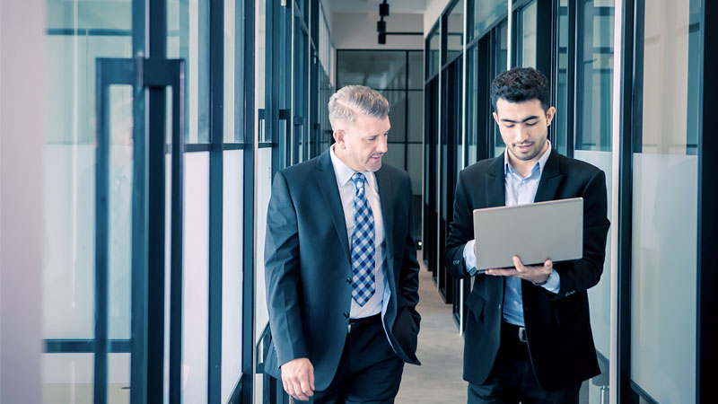Two individuals in suits walk as they work on a laptop