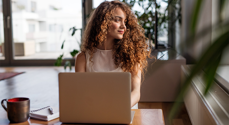 A person with curly hair sitting in front of a laptop at a desk with natural light coming from the window