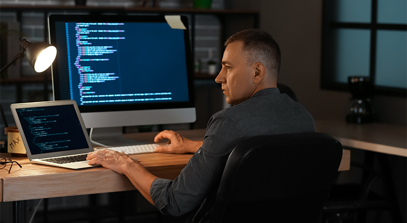 A person sitting at a desk in an office environment, working on a laptop with code on the screen, and a larger monitor with similar code visible