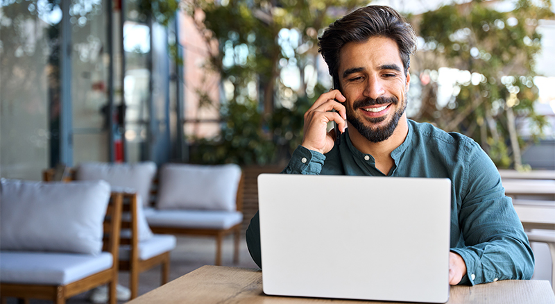 A person sitting at a table in an outdoor environment, working on a laptop while talking on a cellphone