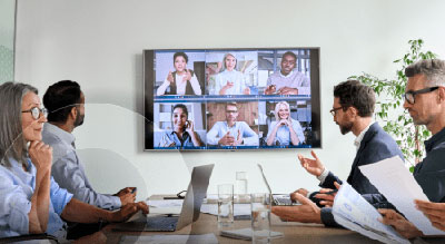 A table of people engage in an online conference, looking at a large screen displaying six people