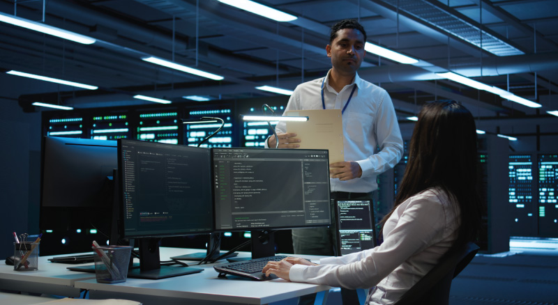A man and woman collaborate in front of a computer screen, engaged in discussion or work-related tasks.