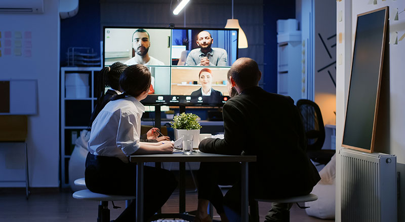 Three people sitting at a table while viewing a meeting with four individuals displayed on a television screen