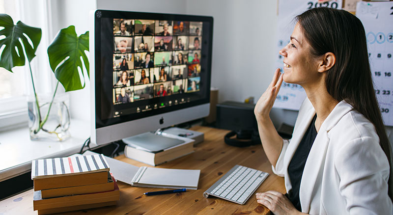 A person waving at a webcam as a monitor displays an online meeting