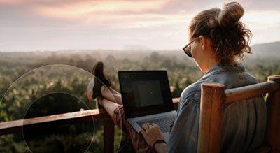 A person working on a laptop outdoors, with feet up on a railing, overlooking a lush landscape at sunset