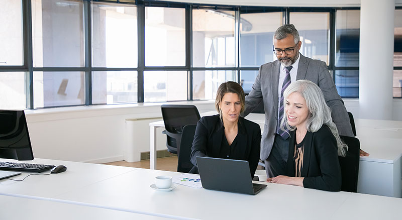 Three professionals in formal attire are gathered around a laptop on a desk in a well-lit modern office