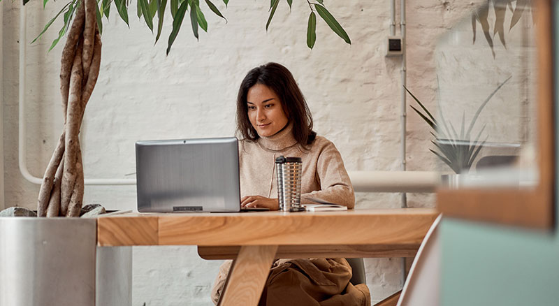 A person is working on a laptop at a wooden table in a room with white brick walls and green plants, under soft natural lighting