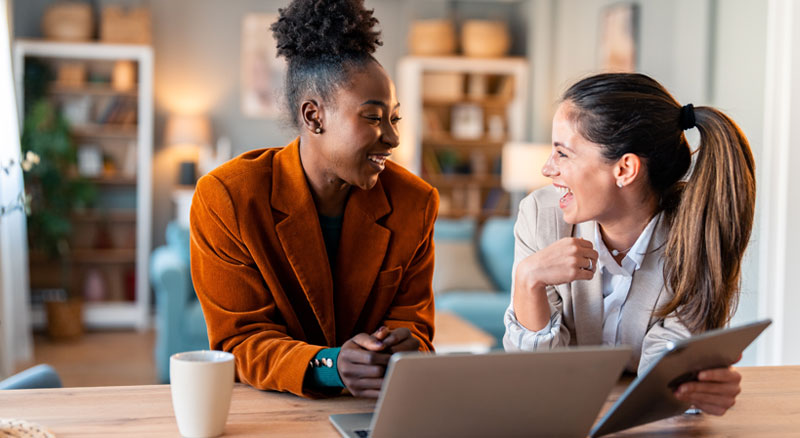 Two smiling individuals talk while using a laptop and tablet