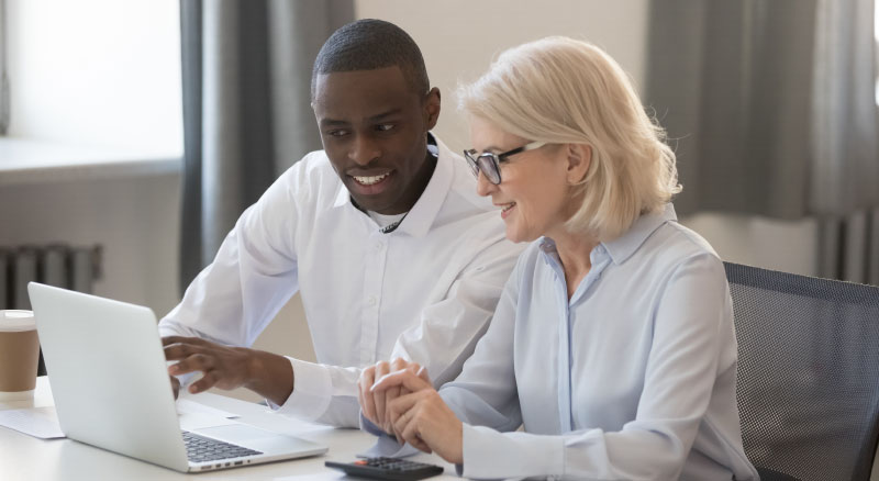 Two individuals are sitting at a table, working together on a laptop in an office setting